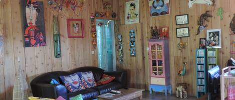 A cozy living room with a wooden wall, a black sofa with colorful cushions, a yellow chair, a wooden coffee table, and various wall decorations including paintings and a bookshelf.