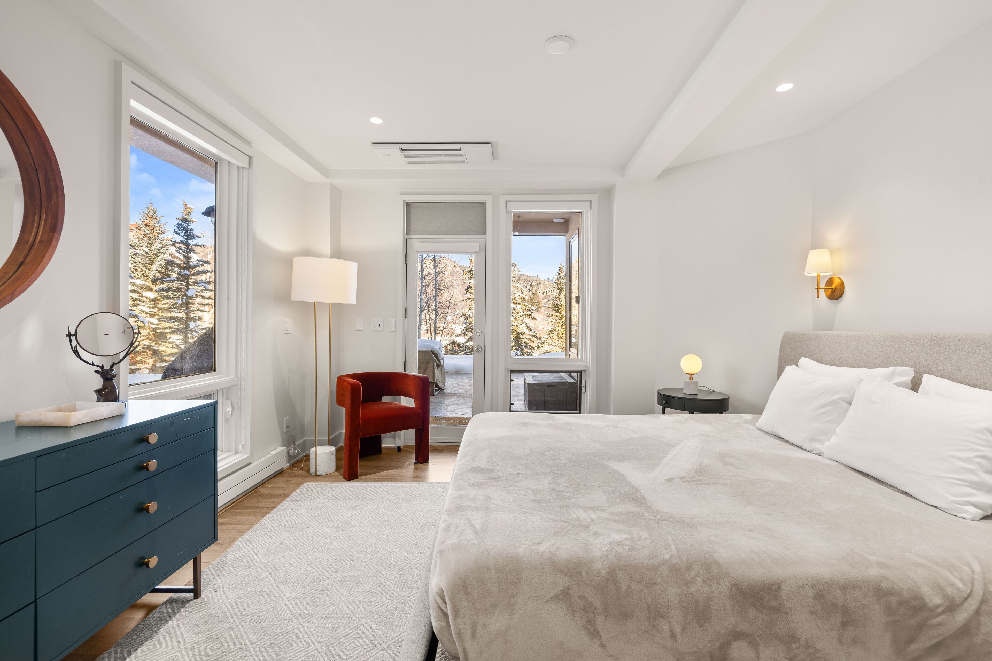 Une chambre à coucher comprenant un lit, une commode, une chaise, une lampe et une fenêtre donnant sur des arbres.