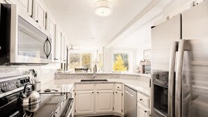 Una cocina moderna con armarios blancos, electrodomésticos de acero inoxidable, encimera de granito y un amplio ventanal con vistas a árboles.
