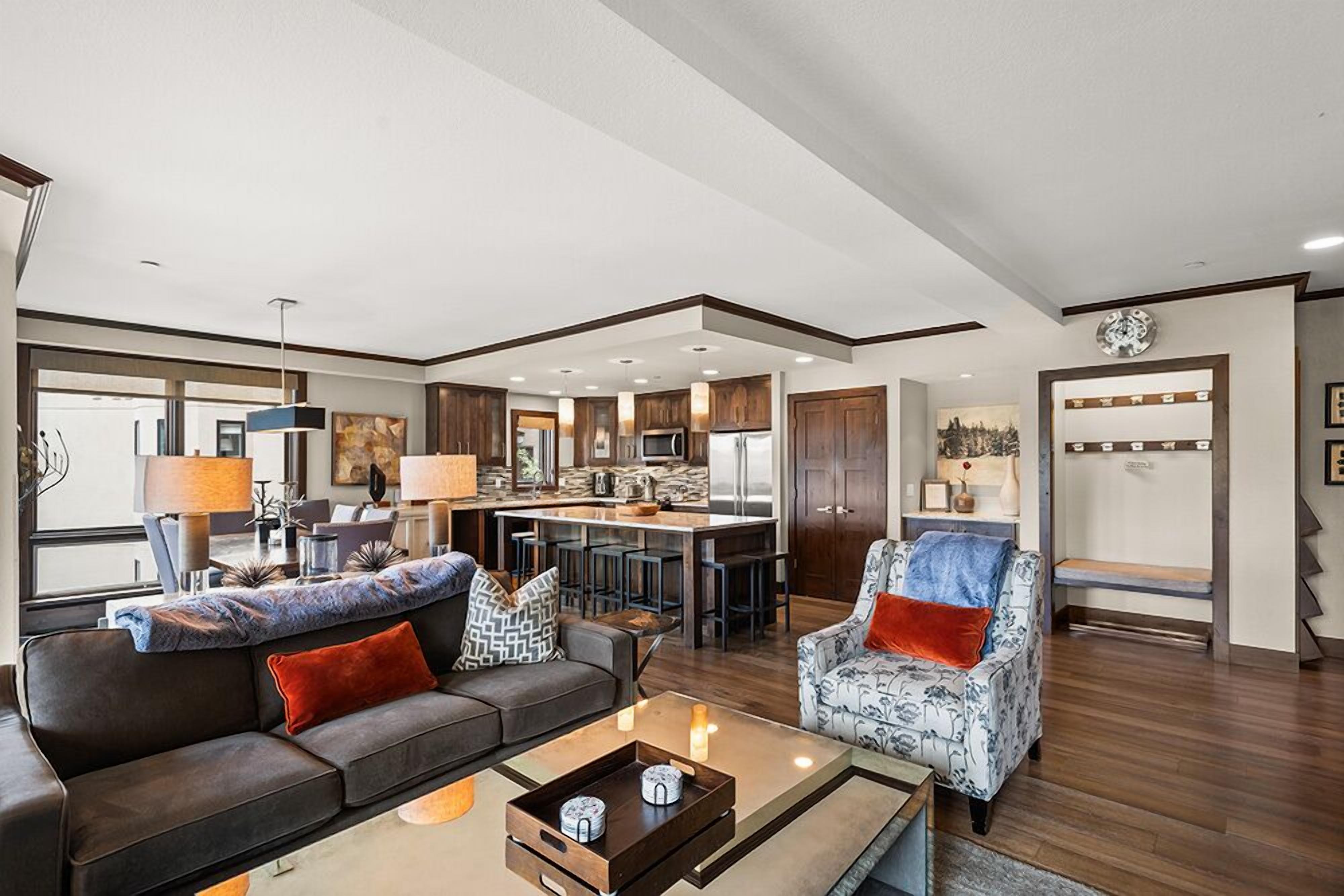 A spacious living room with a sofa, armchair, and coffee table. A kitchen with bar stools is visible in the background.