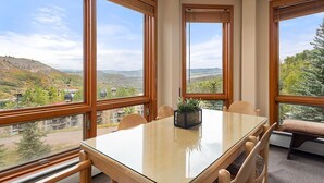 A dining area with a table and chairs, large windows with wooden frames, and a view of a hilly landscape. - Chamonix 37 | Snowmass Ski-In/Ski-Out Condo w/Shared Hot Tub & Pool/Private Washer/Dryer/WiFi (Snowmass Village)