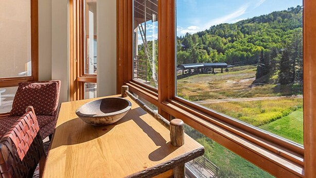 Une table à manger en bois, agrémentée d’un saladier, une chaise à motifs et une vue sur un paysage boisé, visible à travers de grandes fenêtres.