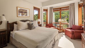 Une chambre spacieuse avec un grand lit, un coin salon avec un canapé et des fauteuils, et une vue sur une forêt à travers de grandes fenêtres.