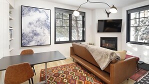 A modern living room with a sofa, a dining table, and chairs. There is a fireplace, a large TV mounted on the wall, and a window with a view of trees. - Christiana Lodge 102A | Studio in Aspen with Hot Tub access (Aspen)