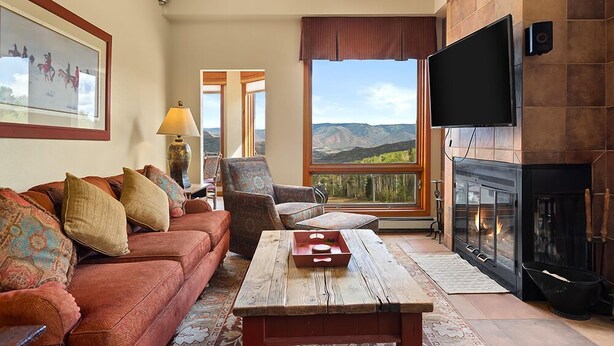 A living room with a sofa, armchair, and a fireplace. There's a large window offering a mountain view. - Chamonix 54 | Snowmass Ski-In/Ski-Out Condo with Pool, Hot Tub, Free WiFi, & Airport Shuttle (Snowmass Village)
