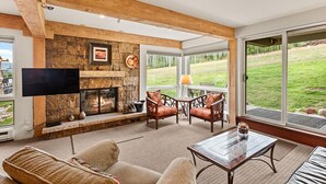 A living room with a stone fireplace, wooden beams, a flat-screen TV, and a view of a grassy yard. - Enclave 102 | Snowmass Ski-In/Ski-Out Condo w/Shared Pool/Hot Tub (Snowmass Village)