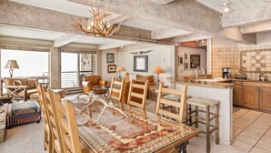 A spacious living area with a large wooden dining table, comfortable seating, and a kitchen with a tiled backsplash. - Enclave 201 | Snowmass Ski-In/Ski-Out Condo w/Shared Pool/Hot Tub/Gym/WiFi (Snowmass Village)