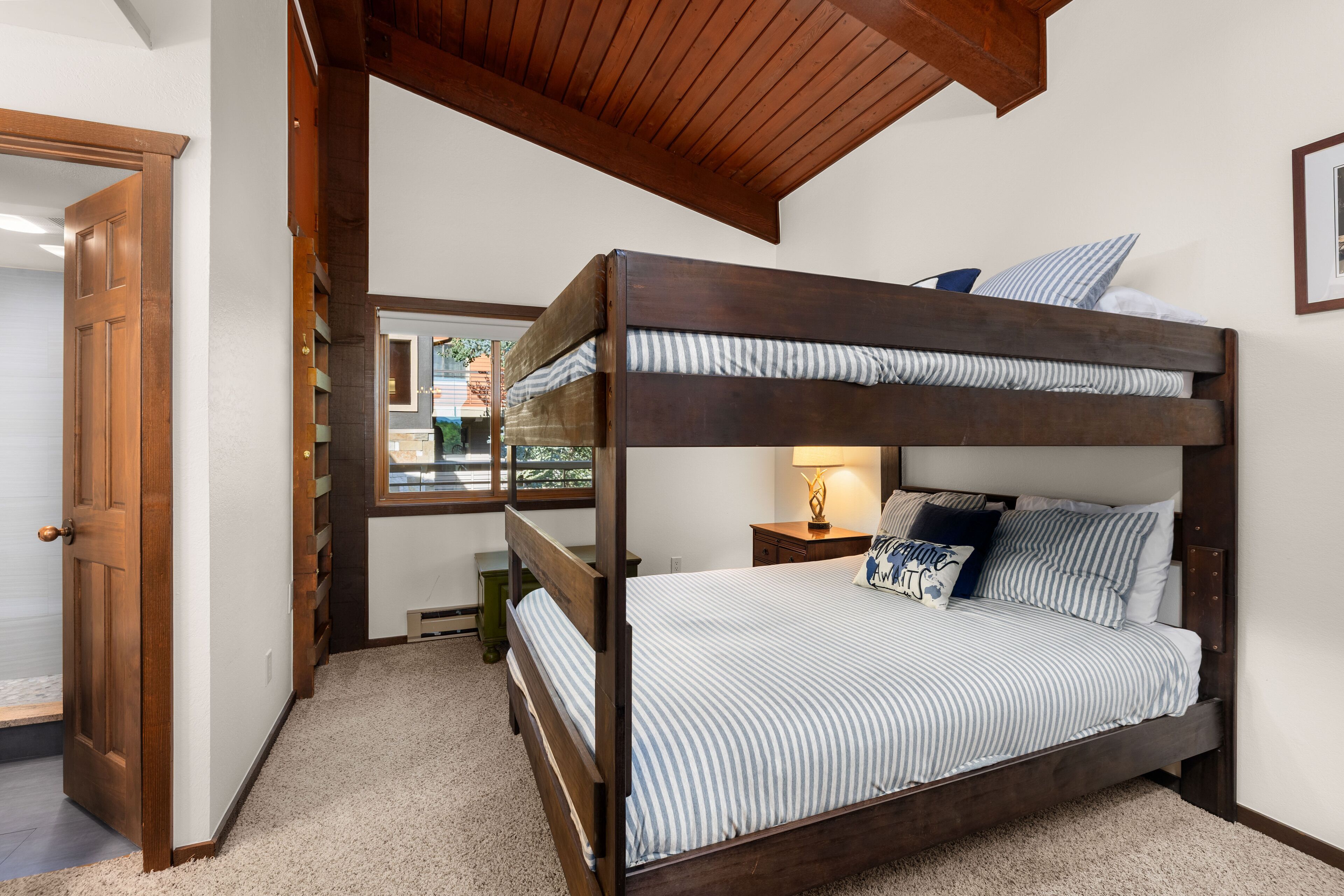 A bunk bed with a striped bedspread and pillows, a wooden ceiling, and a window with a view of greenery.