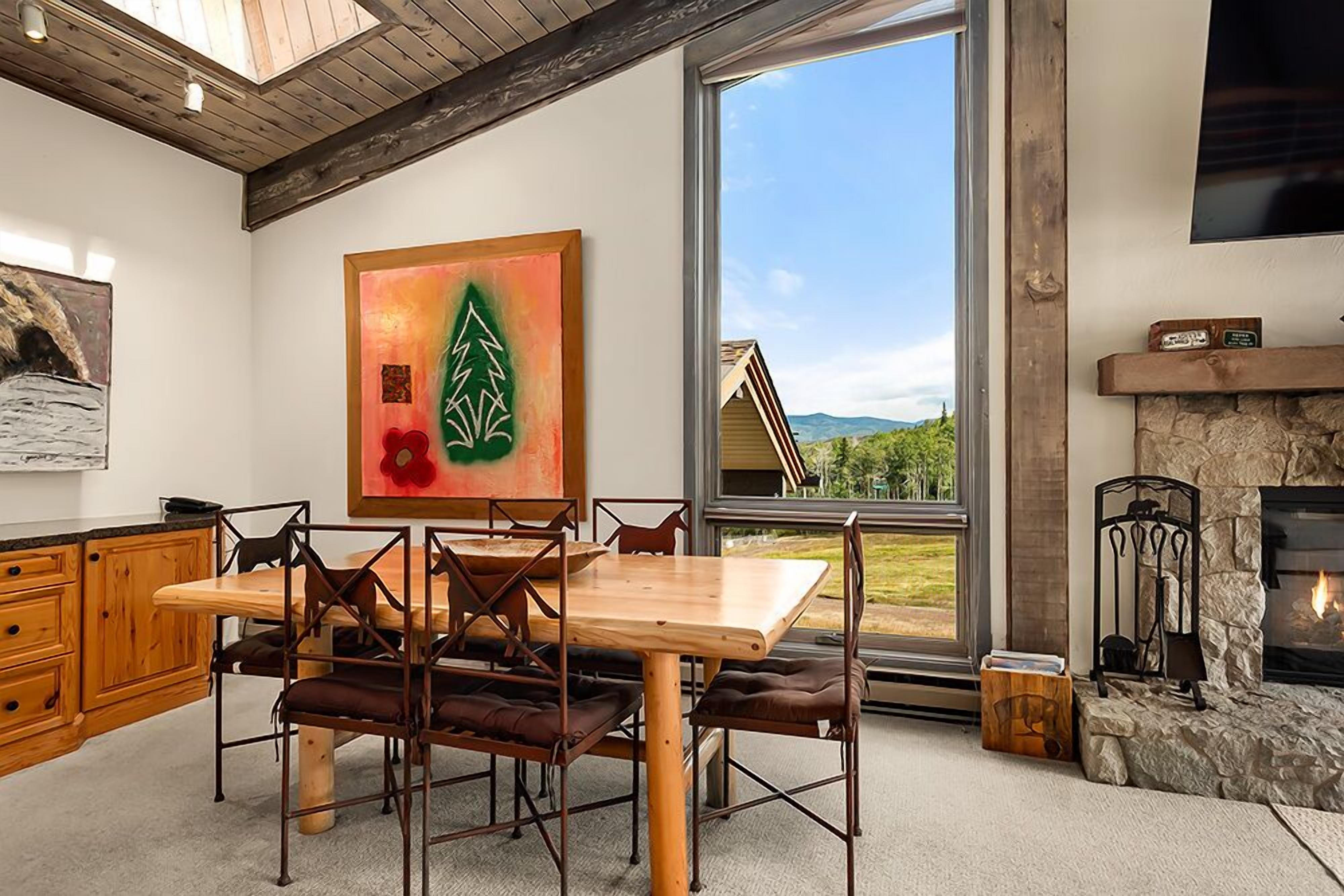 Ein Essbereich mit Holztisch und Stühlen, einem Kamin und einem großen Fenster mit Blick auf ein Haus und Berge.