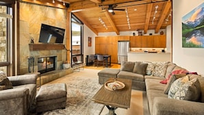 A spacious living room with a stone fireplace, wooden ceiling beams, and a large TV mounted on the wall. - Enclave 312 | Ski-In/Ski-Out Condo, Top Floor w/ Hot Tub, Gym and Out Door Pool in Snowmass (Snowmass Village)