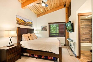 Une chambre avec un plafond en bois, un lit recouvert d’une courtepointe blanche, une table de chevet, un téléviseur fixé au mur et une fenêtre avec des volets.