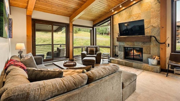 A cozy living room with a stone fireplace, comfortable seating, and large windows overlooking a grassy area. - Enclave 312 | Ski-In/Ski-Out Condo, Top Floor w/ Hot Tub, Gym and Out Door Pool in Snowmass (Snowmass Village)