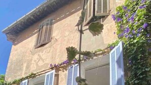 Ein zweistöckiges Gebäude mit einer Steinfassade, mehreren Fenstern mit Fensterläden und einer mit violetten Blumen bewachsenen Mauer.