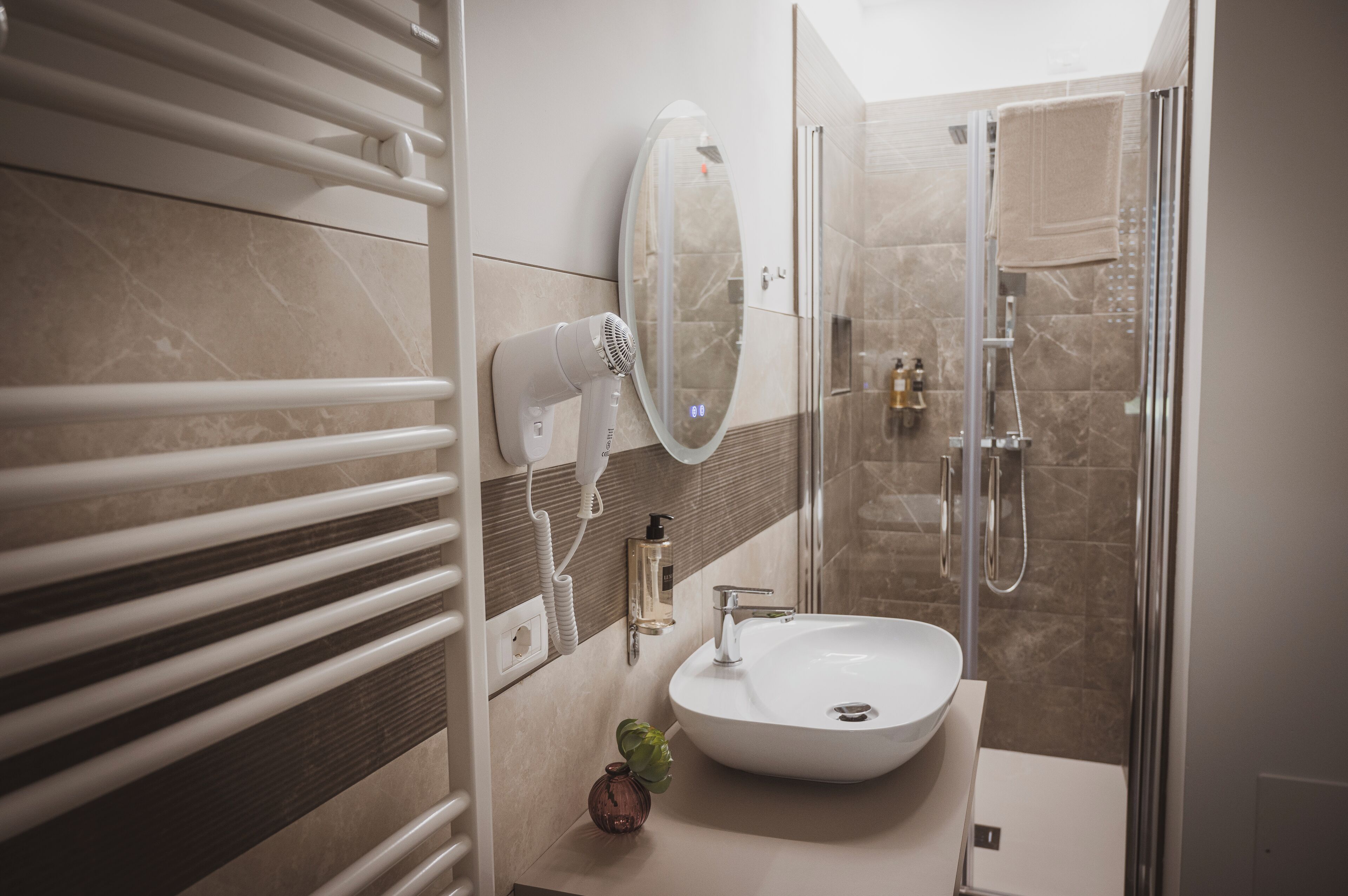 Une salle de bain moderne dotée d’un grand miroir ovale, d’un sèche-cheveux intégré, d’un lavabo et d’une cabine de douche en verre.