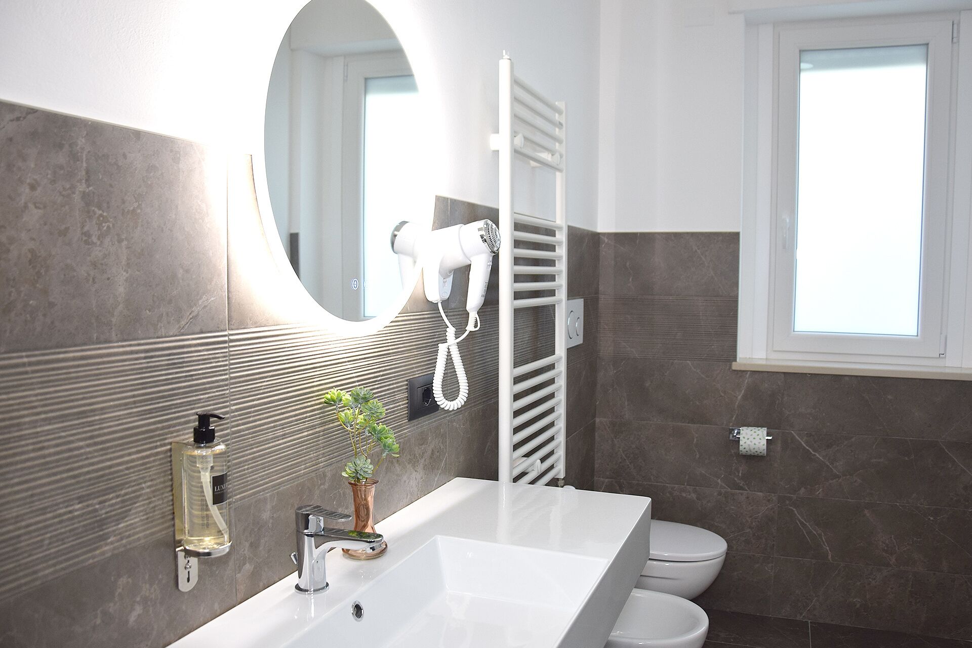 Un baño moderno con un lavamanos blanco, un espejo, un secador de pelo, un perchero para toallas, un inodoro y una ventana.