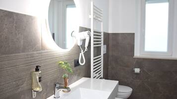 Un baño moderno con un lavamanos blanco, un espejo, un secador de pelo, un perchero para toallas, un inodoro y una ventana.