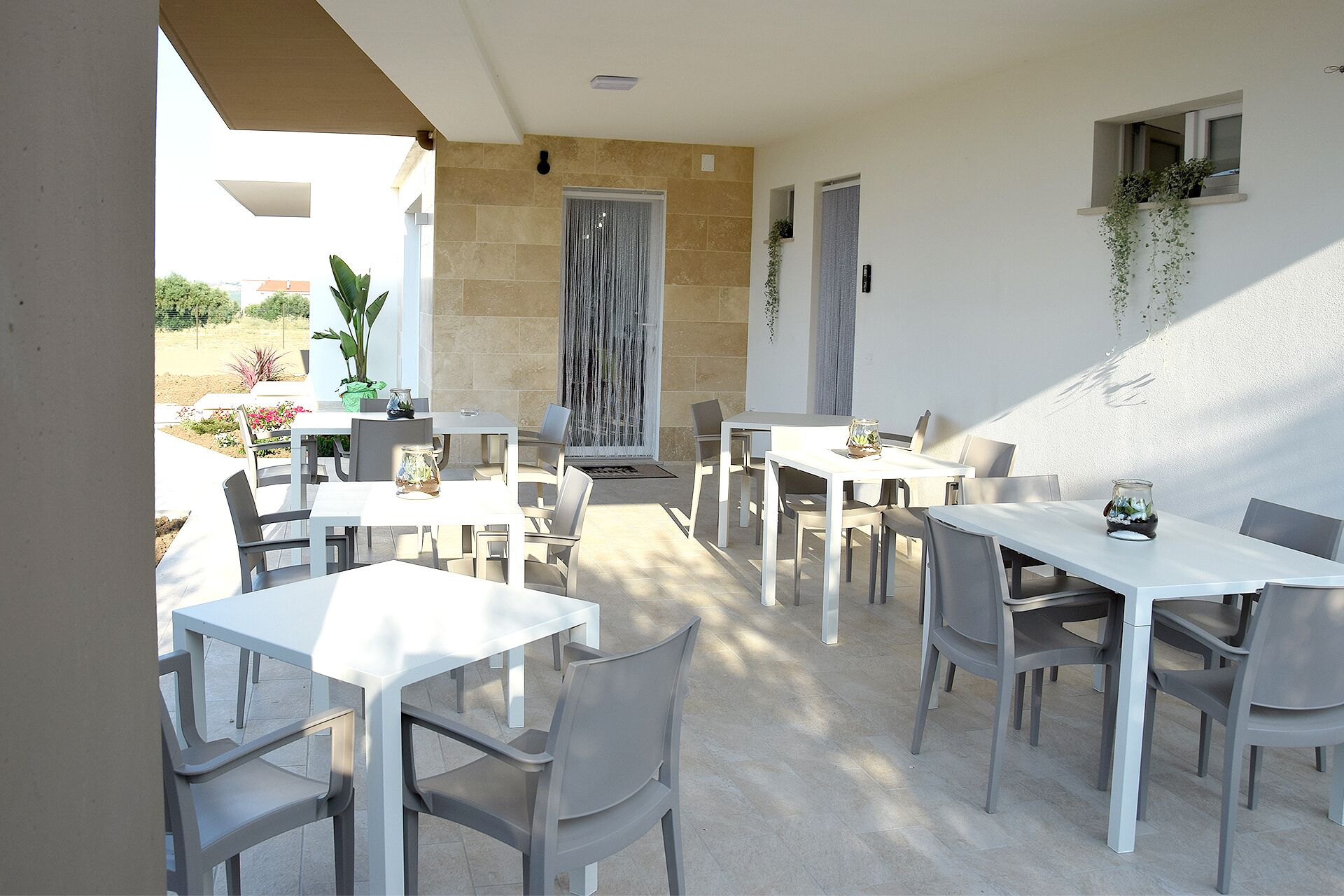 Un espace extérieur aménagé pour les repas, avec plusieurs tables et chaises blanches, des murs en pierre et des plantes en pot.