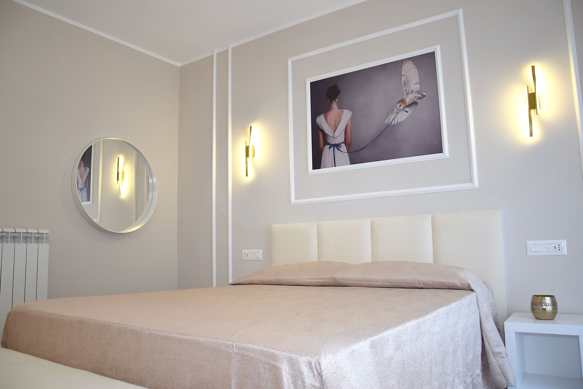 Un dormitorio con cama, un espejo redondo, un cuadro en la pared y dos apliques.