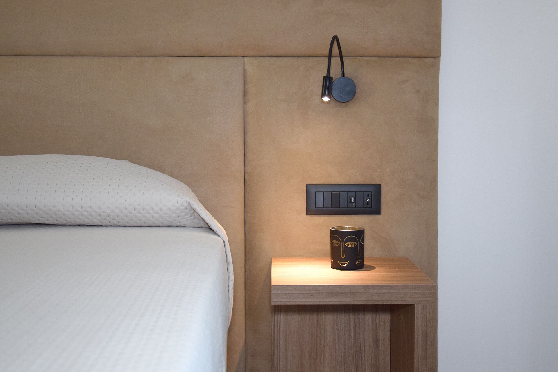 Una cama con cubierta blanca texturizada, una mesita de noche con una lámpara pequeña y un tablero de interruptores en la pared.
