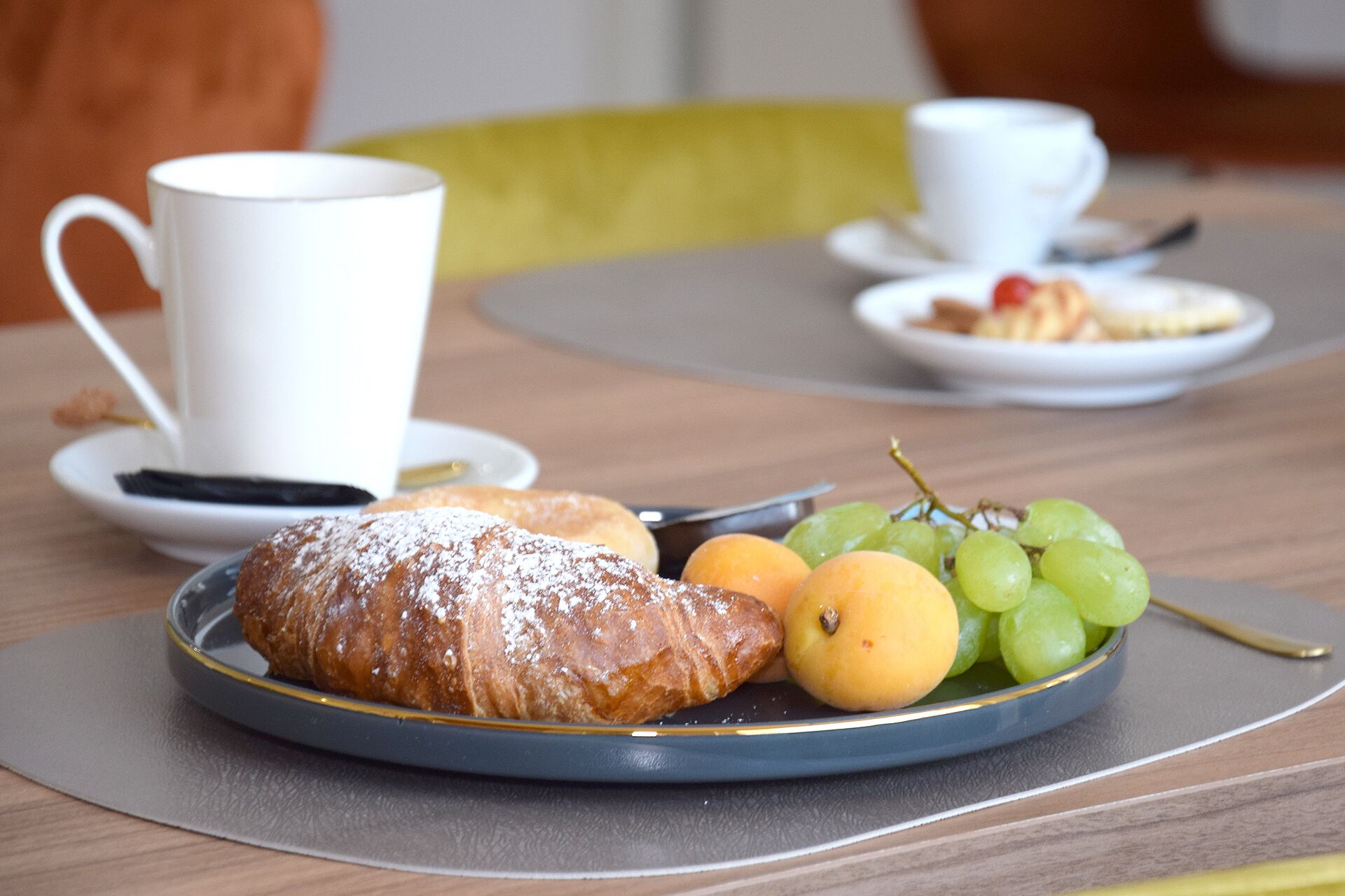 A breakfast setting with a plate of food, including a pastry, grapes, and a lemon, and two white cups on a table.