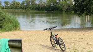 Ein Fahrrad steht auf einem sandigen Strand an einem stillen See, in der Nähe befinden sich eine Bank und ein grünes Tuch.