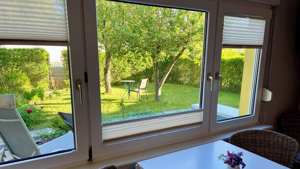 Ein Zimmer mit großem Fenster und Blick auf einen Garten, einem weißen Tisch, Korbstühlen und einer Blumenvase.