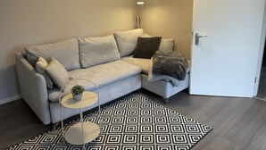 Ein modernes Wohnzimmer mit einem grauen Sofa, einem gemusterten Teppich, einem kleinen Beistelltisch mit einer Pflanze und einer Stehlampe.