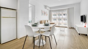 Et moderne køkken med hvide hårde hvidevarer, et spiseområde med et rundt bord og stole, samt et stort vindue med udsigt til bygninger.