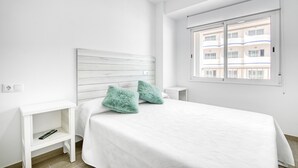 Ein schlicht eingerichtetes Schlafzimmer mit einem weißen Bett, zwei grünen Kissen, einem weißen Nachttisch und einem Fenster mit Blick auf Gebäude.