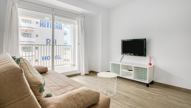 En moderne stue med en beige sofa, et fladskærms-tv monteret på væggen og en skydedør, der fører ud til en balkon.