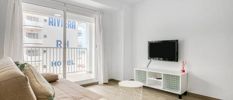 Una sala de estar moderna con un sofá beige, un televisor de pantalla plana montado en la pared y un ventanal corredizo que da a un balcón.
