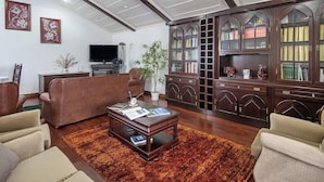A living room with a brown leather sofa, a wooden coffee table with magazines, a television, and a bookshelf.