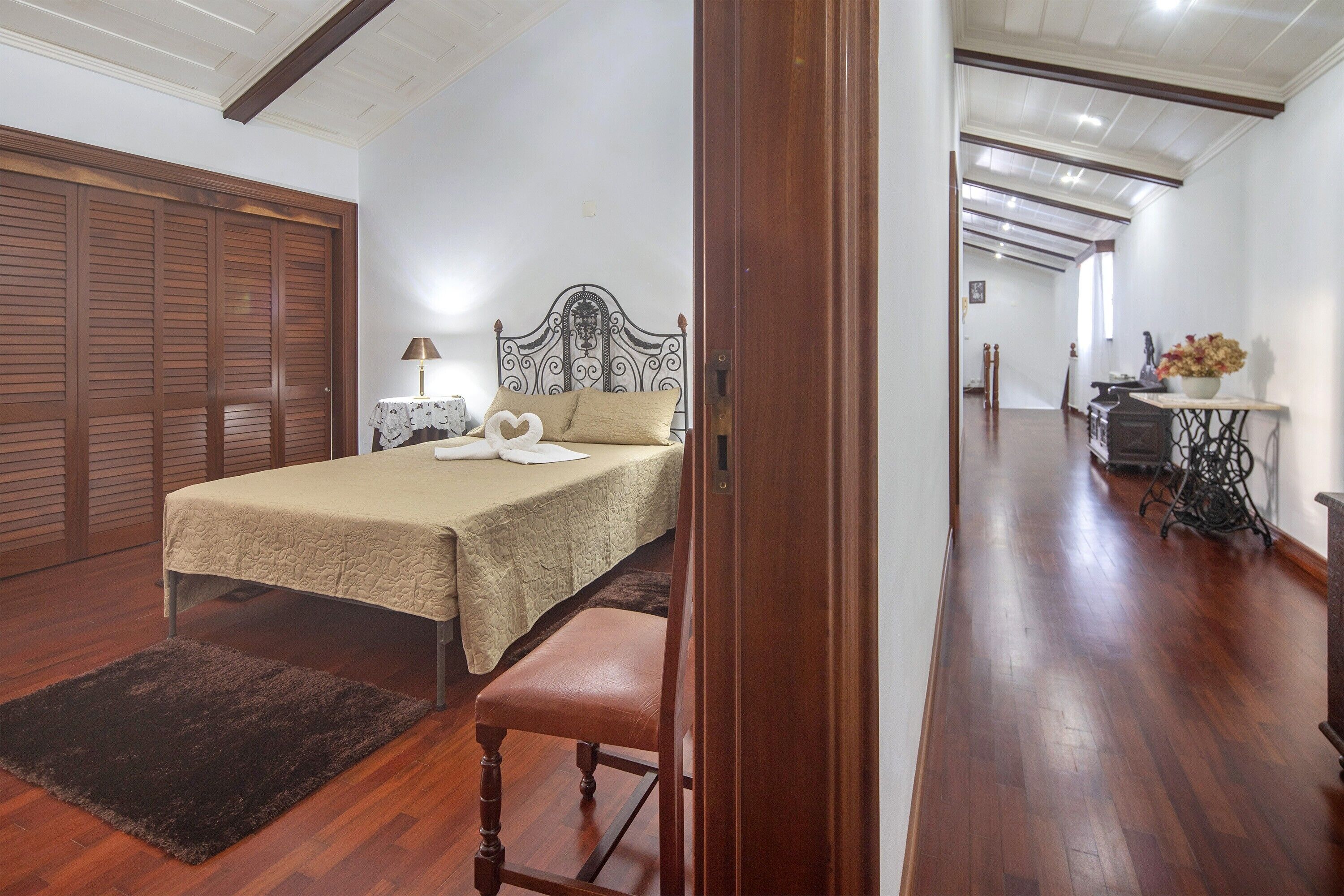 Quarto com cama, um banquinho de madeira, mesa de cabeceira, abajur e cômoda.