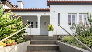 Un bâtiment avec un toit recouvert de tuiles, un escalier menant à une porte marquée « AL », et des plantes en pot disposées autour de l’entrée.