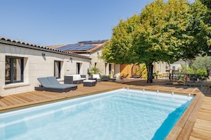 Une piscine entourée de chaises longues et d’une terrasse en bois, le tout bordé d’un bâtiment aux murs en pierre et au toit recouvert de tuiles.
