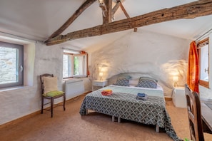 Ein Schlafzimmer mit schrägen Wänden, einem Bett mit gemusterten Betttextilien, einem Sessel mit Kissen und einem Fenster mit Vorhängen.