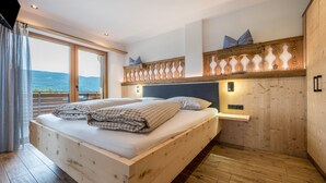 A bedroom with a wooden bed, a nightstand, a wall-mounted light, and a view of the outdoors. - Holiday Apartment "Forchnerhof 4" with Mountain View, 2 Private Terraces & Wi-Fi (Terenten)