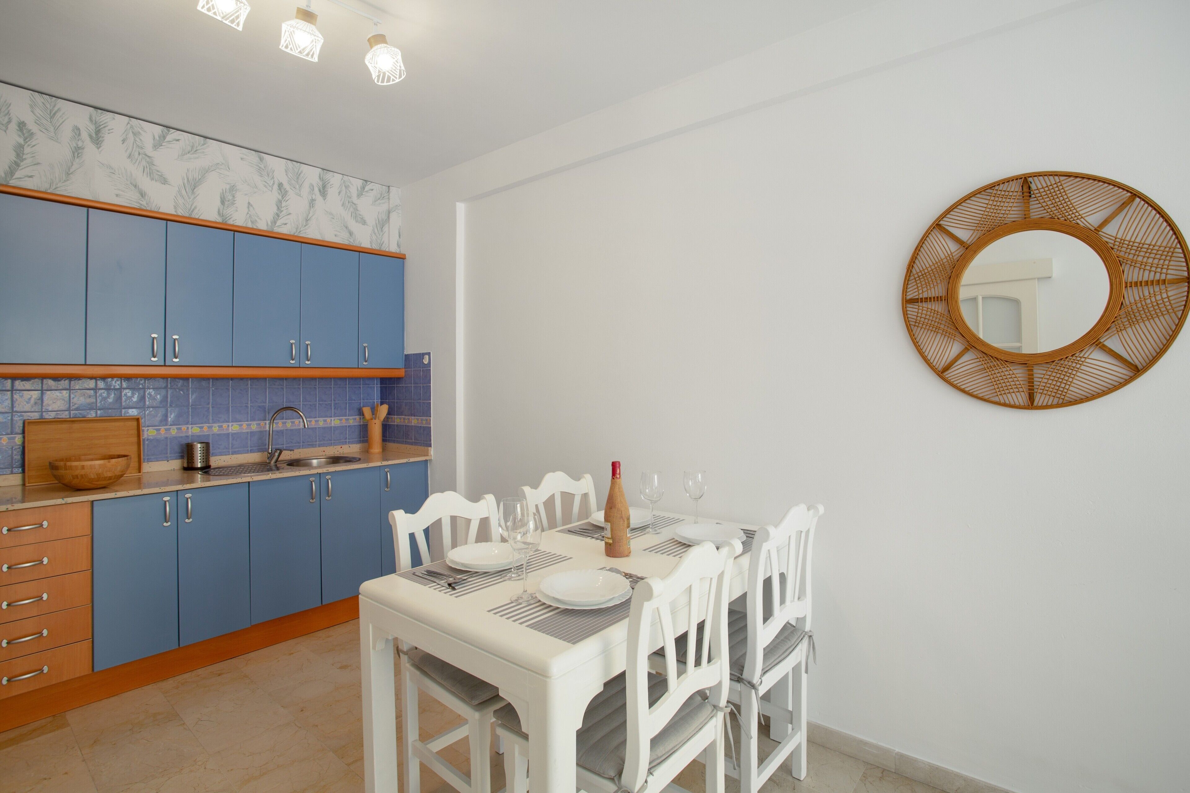 Een keuken met blauwe kastjes, een witte eettafel gedekt met borden en glazen, en een houten kom op het aanrecht.