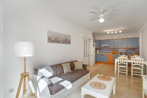 A living room with a sofa, a dining table, and chairs, a kitchen in the background, and a ceiling fan.