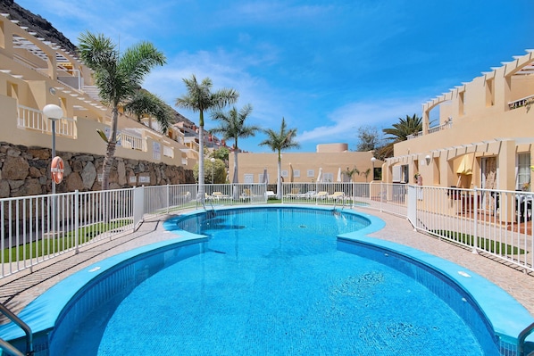 A swimming pool with a curved design, surrounded by a white fence, and adjacent to a building with balconies and palm trees.