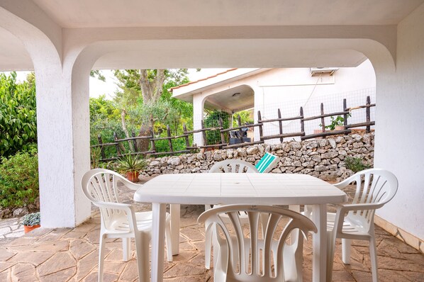 A covered patio with a table and chairs, a stone wall, and a view of a building in the background.