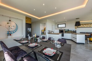 Un espace de salle à manger moderne avec une longue table dressée pour six personnes, une cuisine en arrière-plan et un élément décoratif mural.
