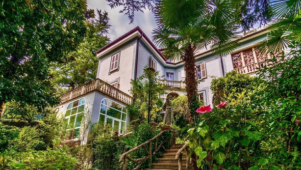 A building with a staircase leading to a balcony, surrounded by lush greenery and a statue.