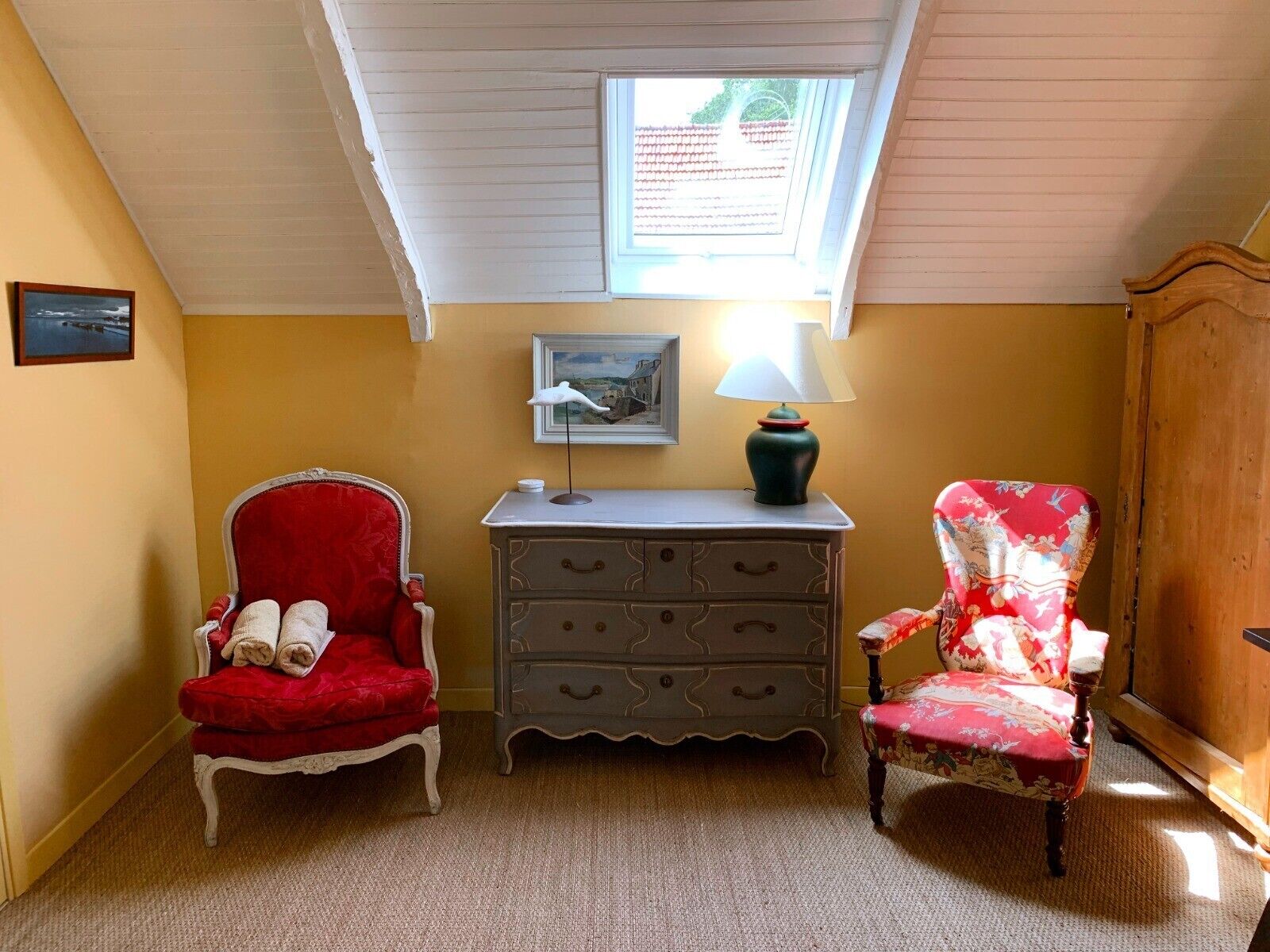 Ein Zimmer mit schräg gedachter Decke, einem Oberlicht, einem roten Stuhl mit zusammengerolltem Handtuch, einer grauen Kommode und einem floralen Sessel.