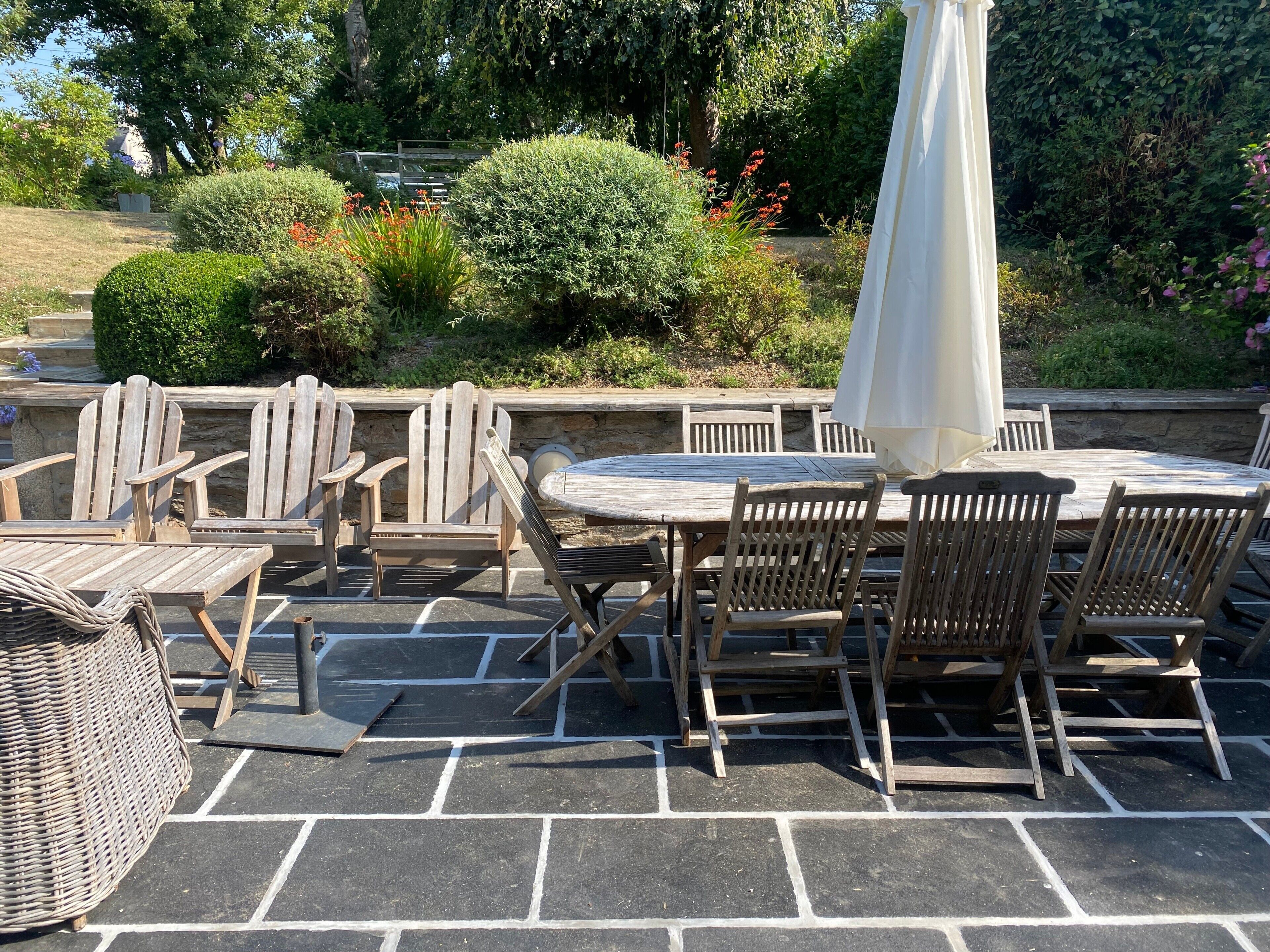 Un patio al aire libre con una mesa redonda y sillas de madera, rodeado de pavimento de piedra y follaje.