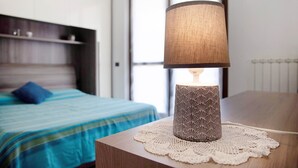 Una cama con colcha azul y verde a rayas, una mesita de noche con una lámpara estampada, y una ventana con persianas al fondo.