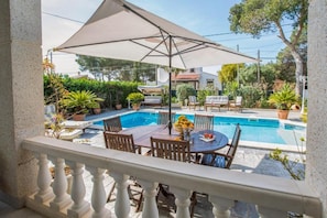 Un área junto a la piscina con una mesa y sillas, una sombrilla grande y plantas en macetas.
