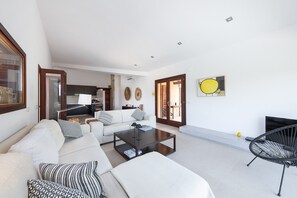 A modern living room with a sofa, coffee table, and a black chair. There is a kitchen in the background, a door leading to an outdoor area, and a painting on the wall.