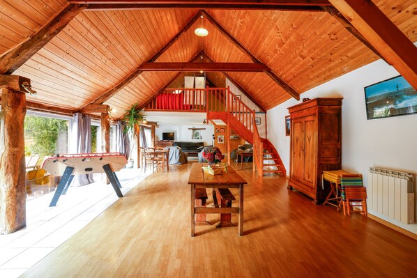 A spacious loft with a high ceiling, wooden beams, a dining area, a staircase, and a view of the outdoors.
