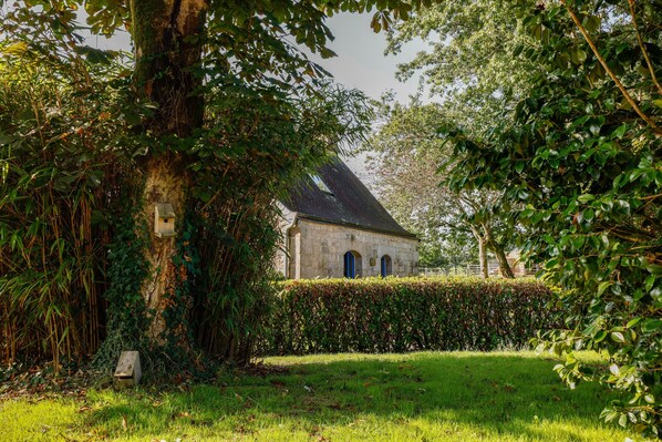 Een gebouw van natuursteen met een schuin dak, omgeven door weelderige begroeiing en een verzorgde haag.